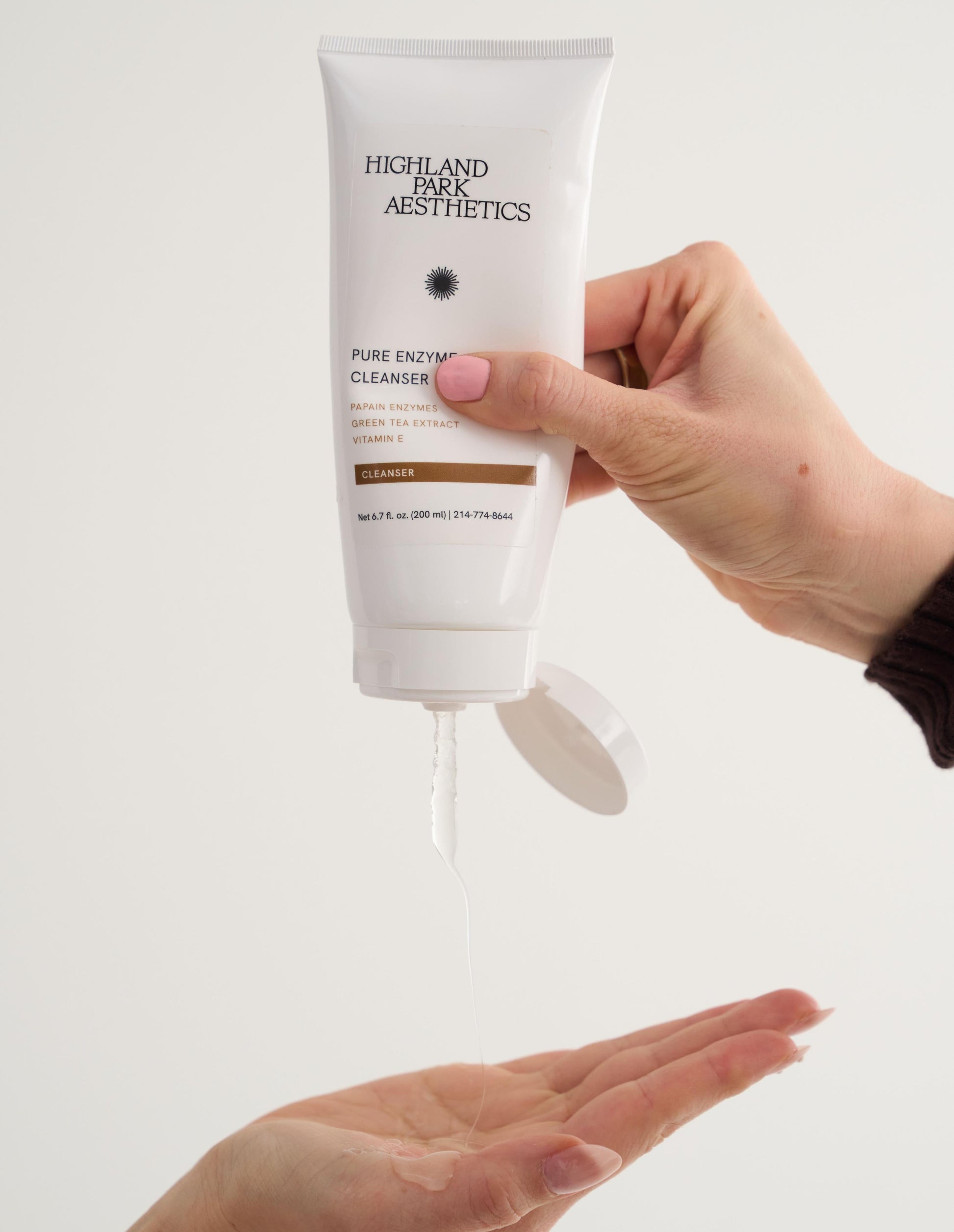 Hand holding a tube of Highland Aesthetics Pure Enzyme Cleanser with another hand below to receive the product.